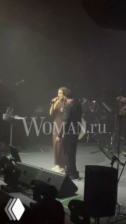 Эмре Алтуг на сцене: певец поёт в микрофон, видна часть оркестра и тёмная сцена, крупный водяной знак Woman.ru