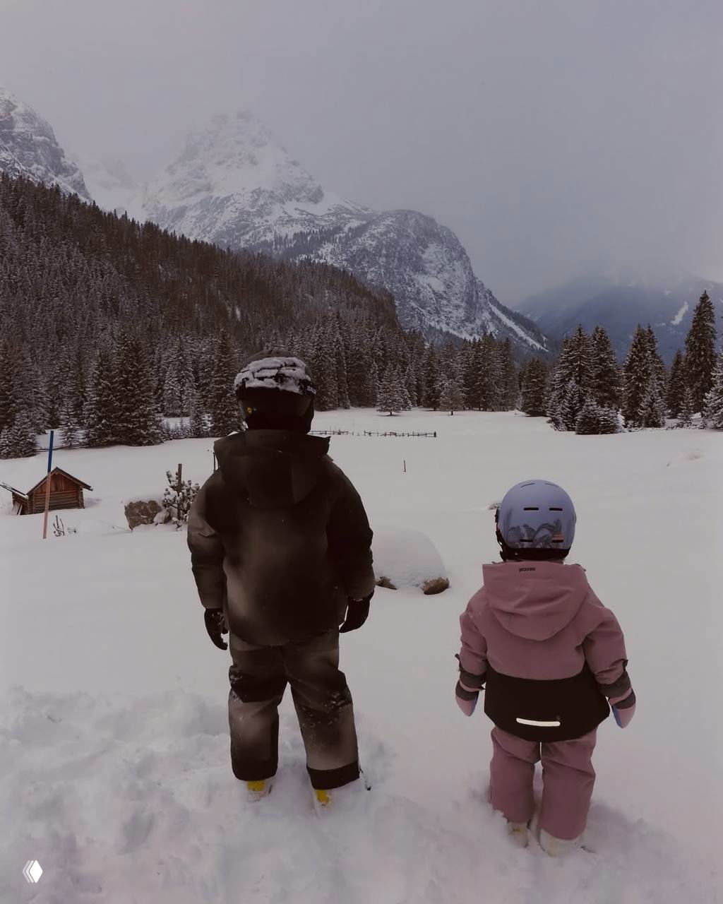 Двое детей смотрят на просторную заснеженную долину и горы, вид сзади подчёркивает масштаб пейзажа и семейный отдых на природе.
