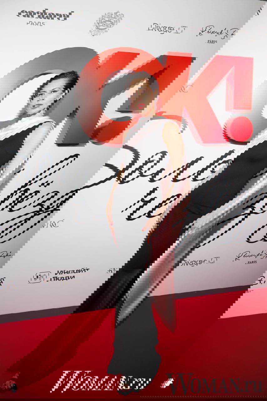 Женщина в облегающем черном платье с драпировкой и розовой накидкой позирует на красной дорожке у логотипа OK!, акцент на силуэте.