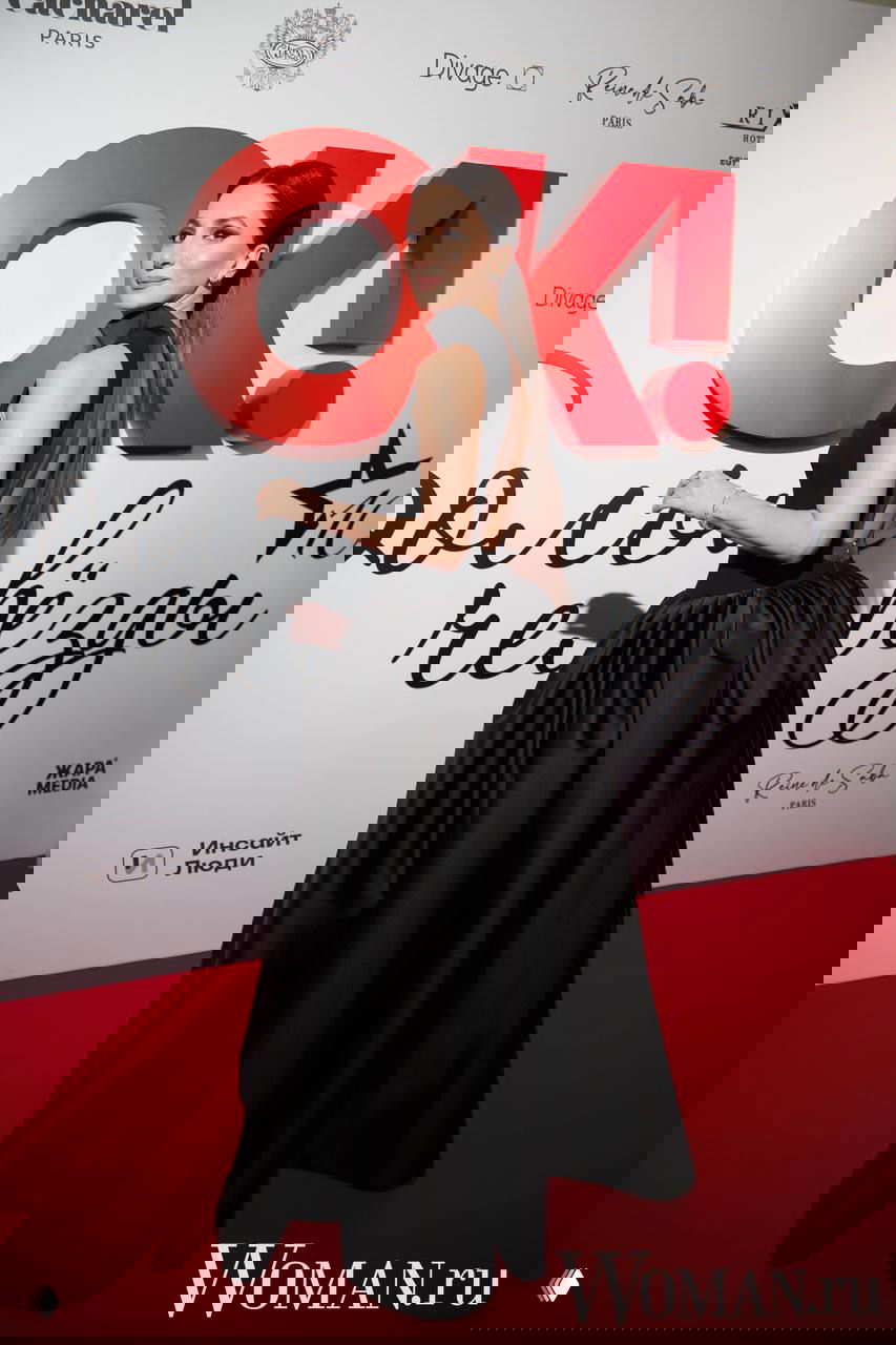 Женщина в закрытом черном платье с открытой спиной и объемной юбкой стоит у брендированного баннера OK!, видна прическа и силуэт платья.