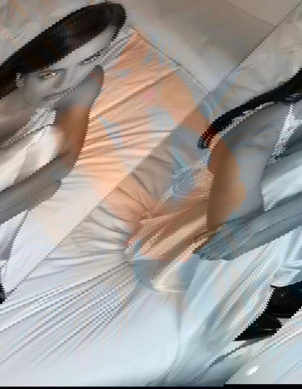 Крупный кадр модели в белом платье в сидячей позе на кровати; внимание на руках, плечах и отражении сатина ткани.