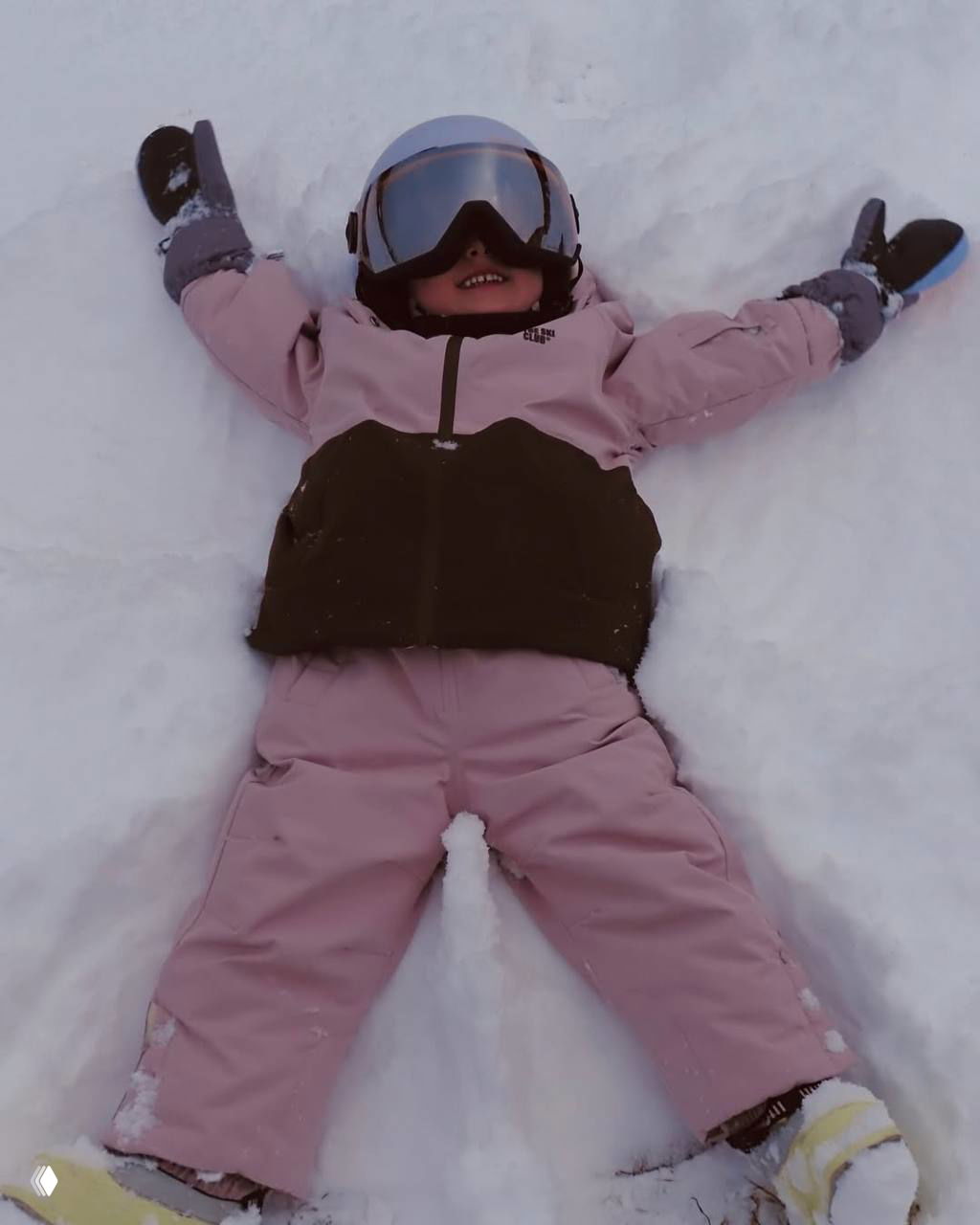 Ребёнок в розовом зимнем костюме лежит в снегу и улыбается, делает «снежного ангела» — игривый момент детского отдыха на морозе.