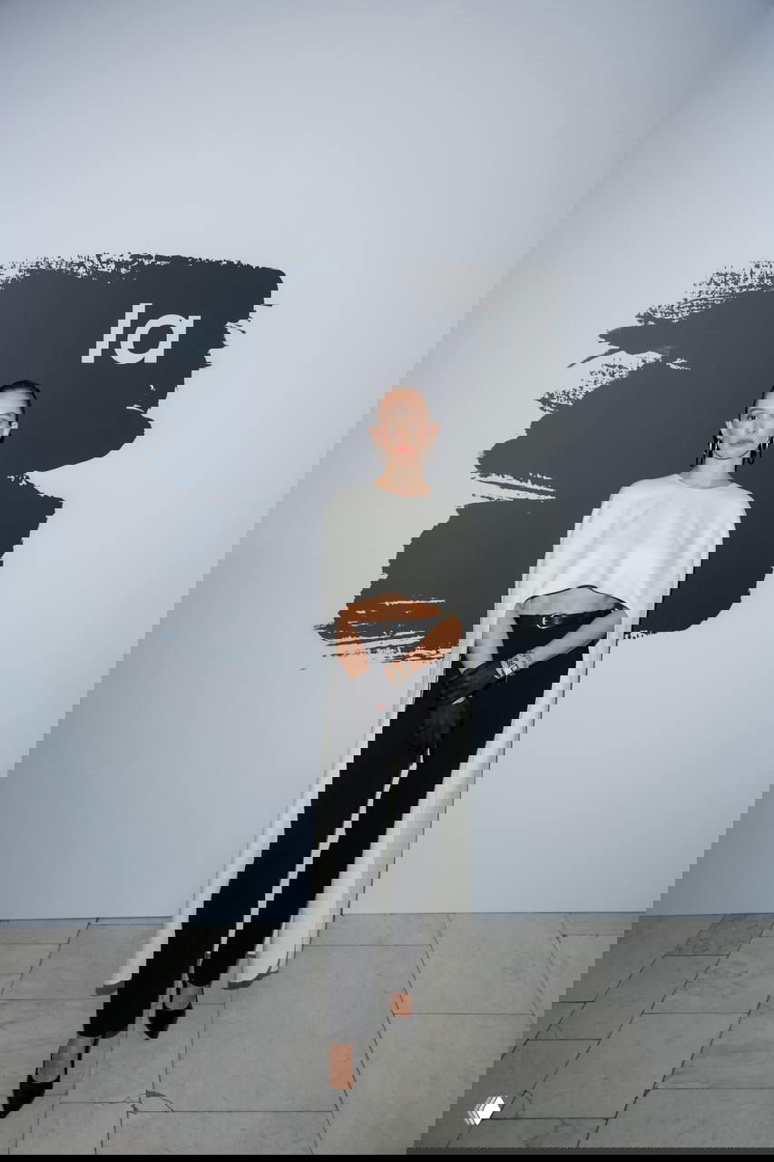 Женщина в белом кейпе и чёрных брюках позирует на фоне графического баннера «la» в зале Третьяковской галереи на событии Lamoda.