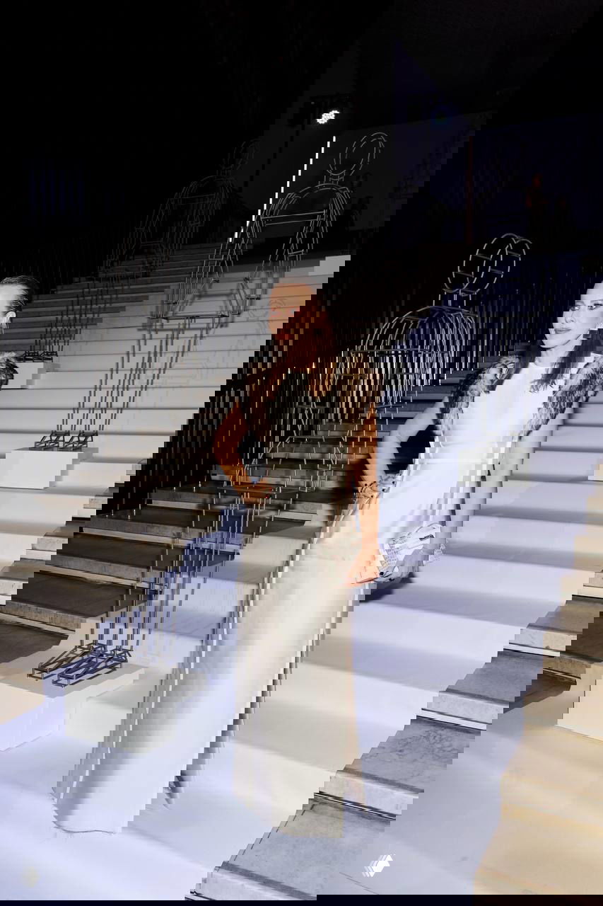 Женщина в светлом длинном платье с меховыми деталями стоит на лестнице выставочного зала — атмосферный снимок с арт‑инсталляцией на фоне.