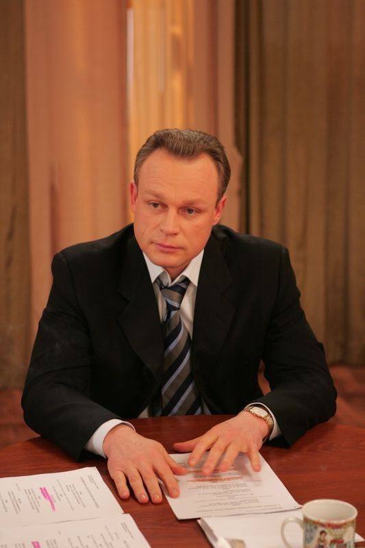 Портретный кадр персонажа в деловом костюме за столом с документами и кружкой, сцена демонстрирует сосредоточенность и рабочую атмосферу в интерьере офиса.