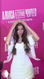 Женщина в белом платье и легкой накидке на красной дорожке перед розовым баннером с надписью «Алиса в стране чудес» на премьере.