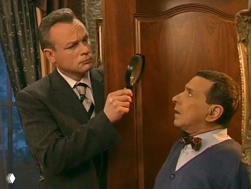 Комедийная сцена у двери: один персонаж рассматривает что-то через лупу, другой выглядит удивлённым; кадр показывает бытовой юмор и экспрессию актёров.