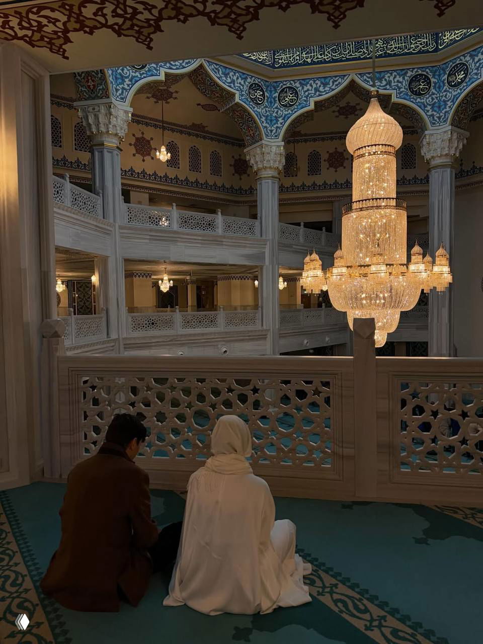 Внутреннее убранство мечети с большой хрустальной люстрой; пара сидит на ковре спиной к камере, видна декоративная резьба и балюстрада.