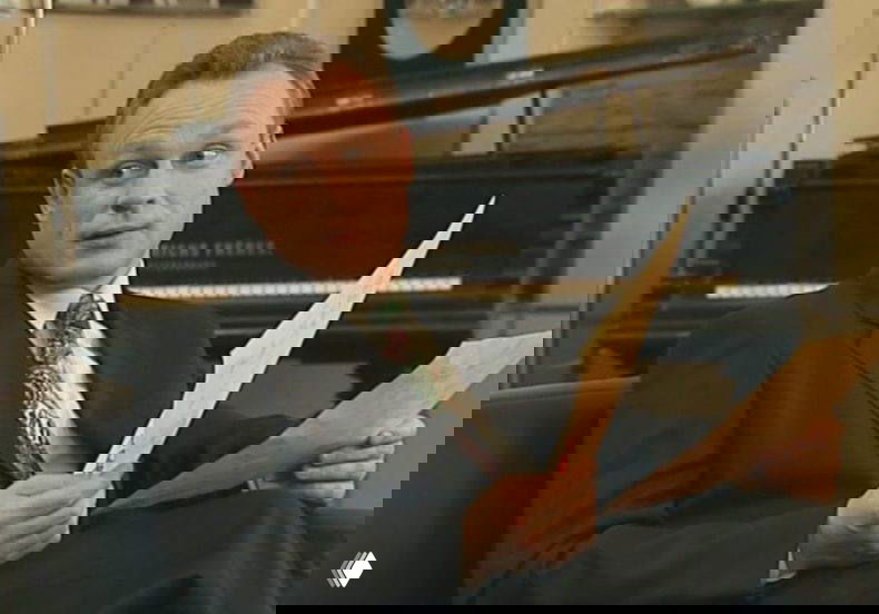 Персонаж в костюме держит документы в руках на фоне пианино; кадр передаёт деловую атмосферу и лёгкую ироничность сцены в телевизионном шоу.