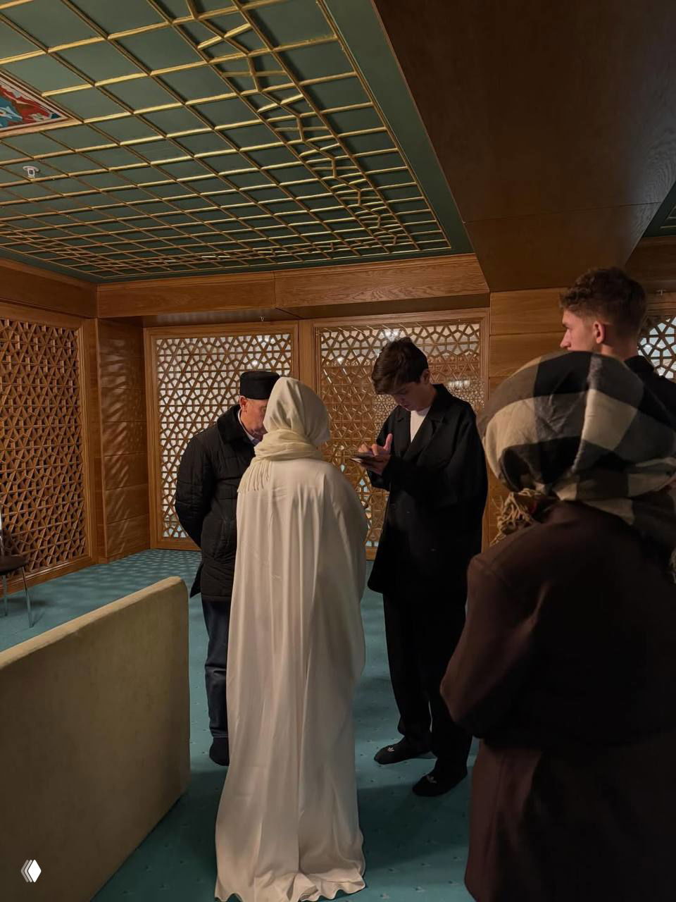 Сцена брачного обряда: невеста в белом платке рядом с мужчиной, вокруг несколько людей и резные деревянные перегородки в помещении мечети.