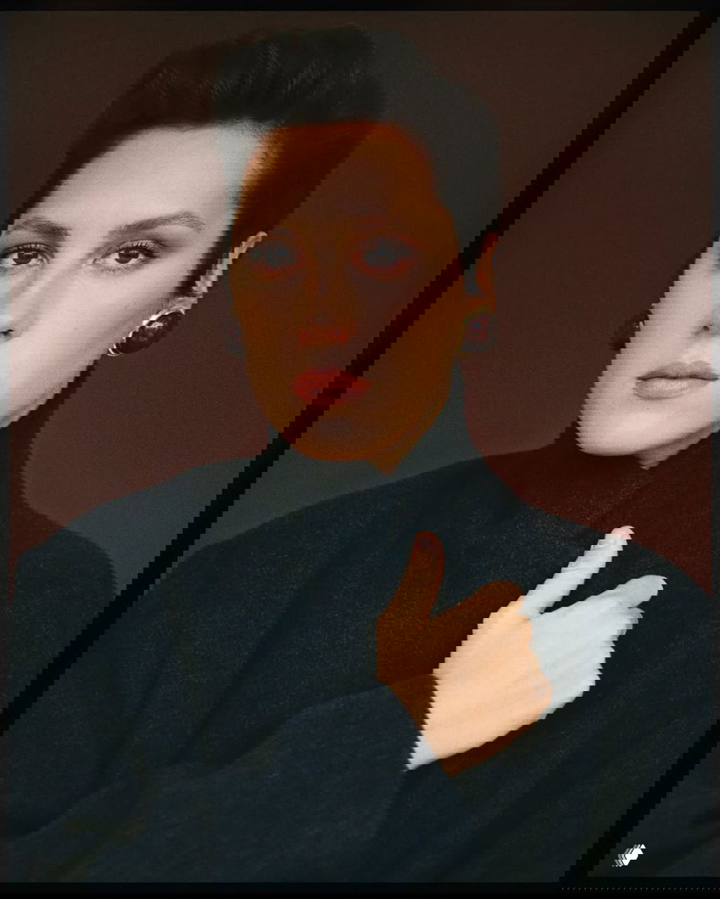 Крупный портрет женщины в тёмном пальто на нейтральном фоне; крупные серьги, естественный макияж и сдержанный элегантный образ.