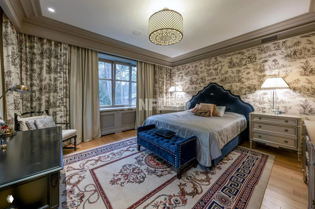 Ещё одна спальня с двуспальной кроватью, бархатным изголовьем, узорной обивкой стен и классическим ковром на деревянном полу; интерьер в светлых тонах.