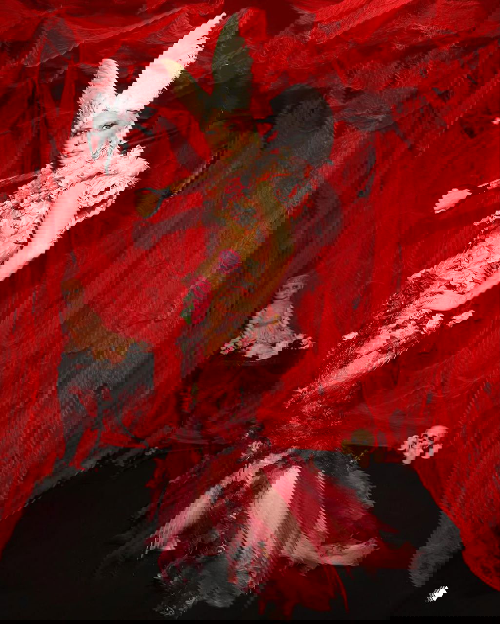 Леди Гага в образе «садовница из Эдема»: сложный цветочный наряд с белыми рогами, драматичная красная декорация, кукла и череп в окружении.