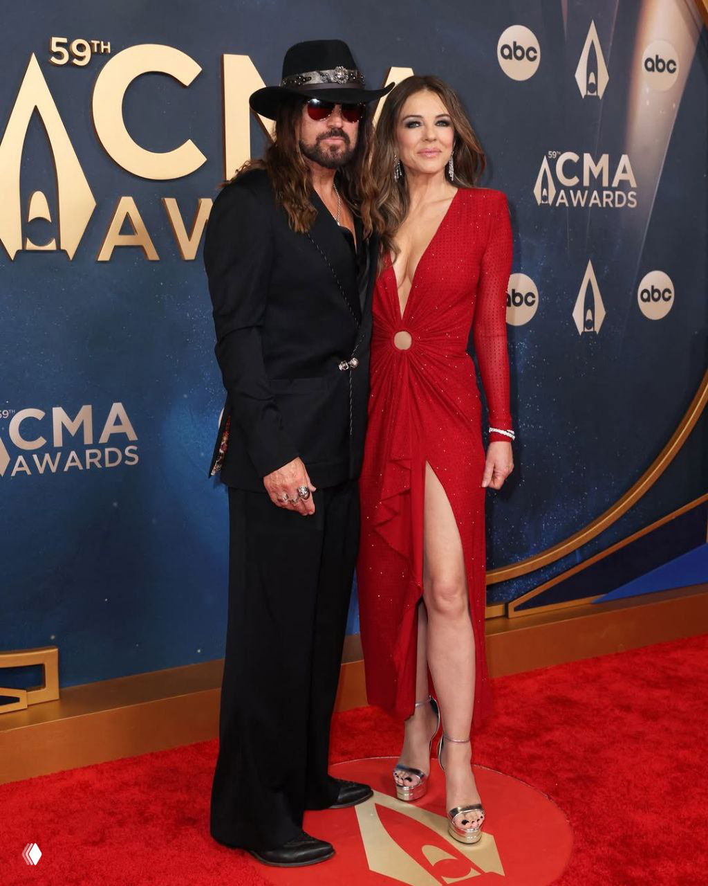 Элизабет Хёрли в красном платье с глубоким декольте и Билли Рэй Сайрус в чёрной шляпе и костюме позируют вместе на красной дорожке CMA Awards, улыбаются в камеру.