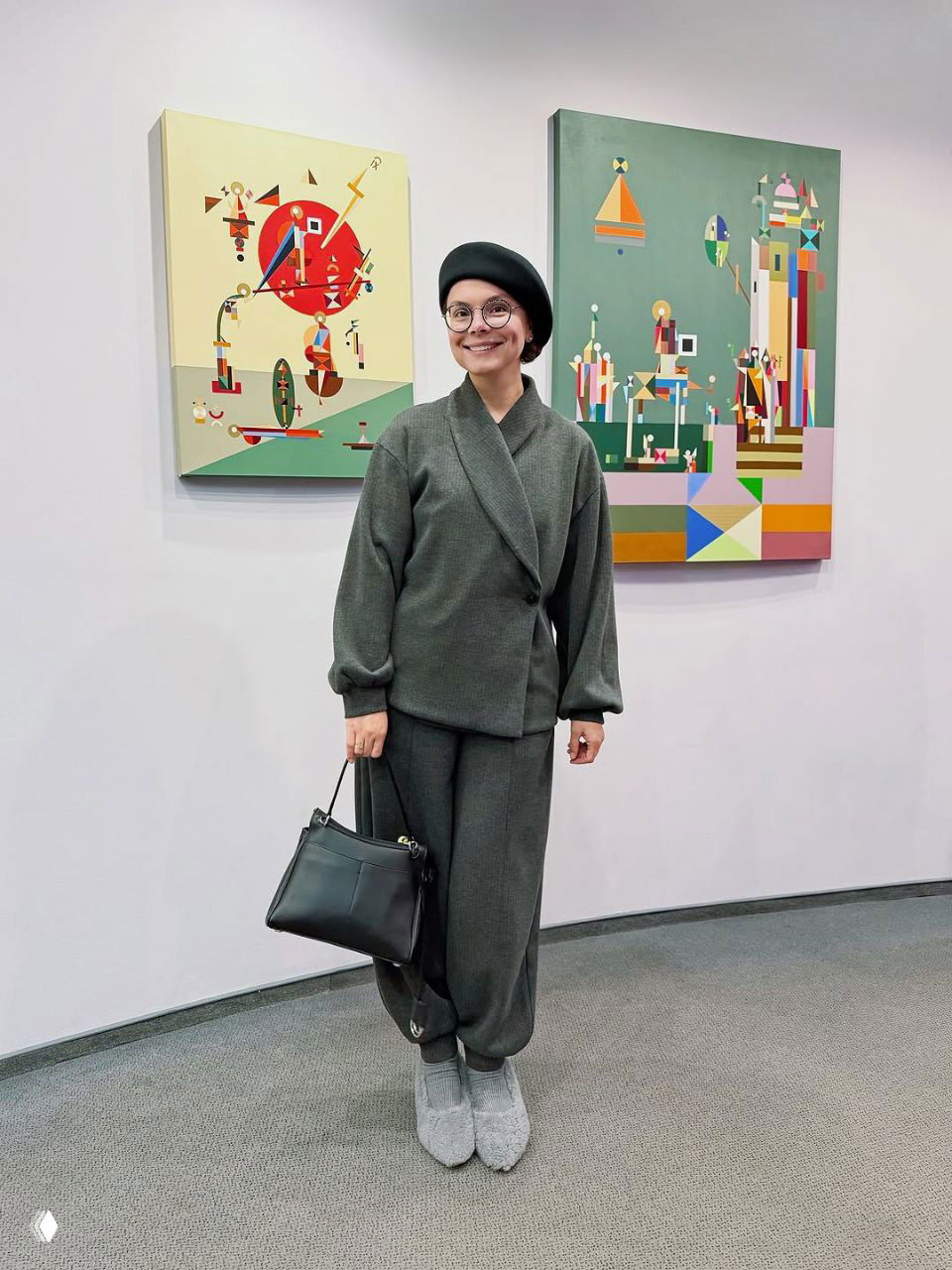 Татьяна Брухунова в тёплом сдержанном серо‑зеленом костюме с беретом и сумкой, позирует в интерьере с яркими картинами на стене.