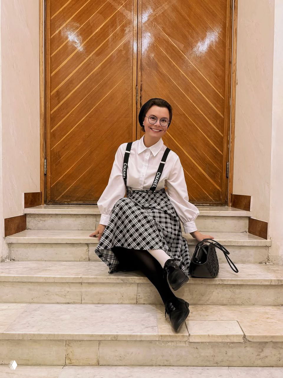 Татьяна Брухунова в белой блузе и клетчатой юбке с подтяжками сидит на ступенях — образ в стиле ретро и школьной эстетики.