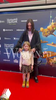 Женщина и маленькая девочка на красной дорожке перед промо-баннером с логотипами спонсоров и постером фильма — кадр с премьеры.