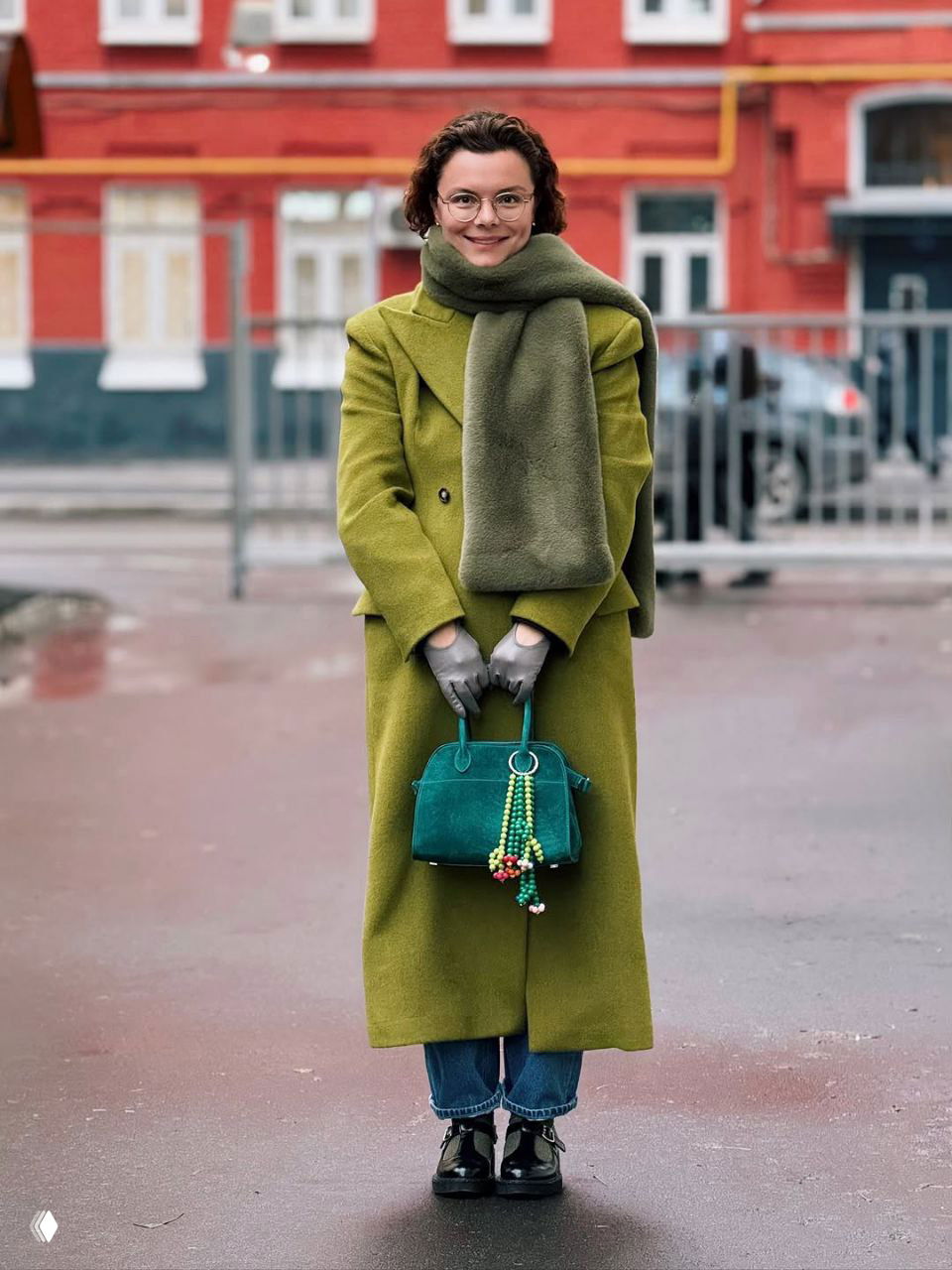 Татьяна Брухунова в ярком зелёном длинном пальто и объёмном шарфе держит бирюзовую сумку с аксессуарами на фоне городского фасада.