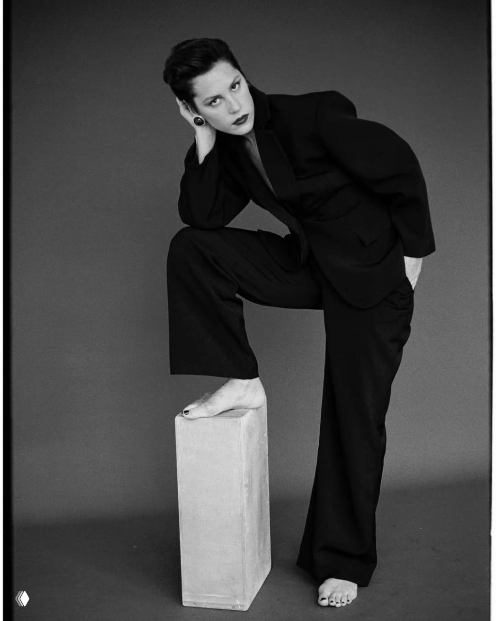 Чёрно‑белая студийная фотография женщины в строгом костюме, динамичная поза с одной ногой на подставке и выразительным взглядом.