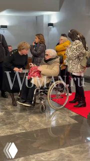 Светлана Крючкова в инвалидной коляске среди гостей в фойе, рядом люди общаются, фотографы и красная дорожка видны на заднем плане.