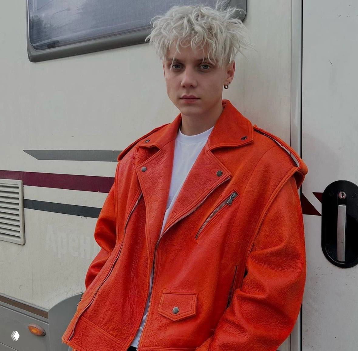 Портрет молодого мужчины с платиновыми кудрявыми волосами в яркой оранжевой кожаной куртке на фоне трейлера — стильный образ артиста.
