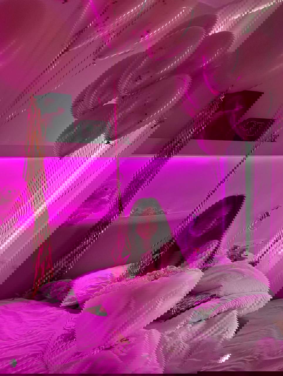 Комната с розовой подсветкой: девушка сидит в постели среди шаров и праздничного декора, атмосферный домашний кадр.