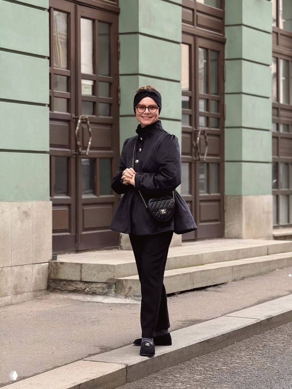 Татьяна Брухунова в чёрном укороченном пальто и чалме стоит у зданий в городской среде, сумка‑плечо и элегантные туфли дополняют образ.