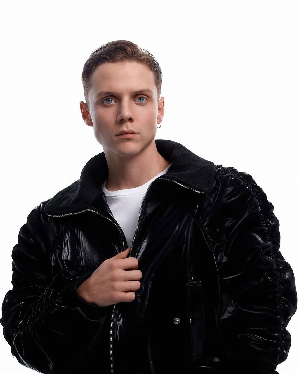 Студийный портрет мужчины в глянцевой чёрной куртке на светлом фоне, серьга в ухе и серьёзный взгляд прямо в камеру — модная фотосессия.