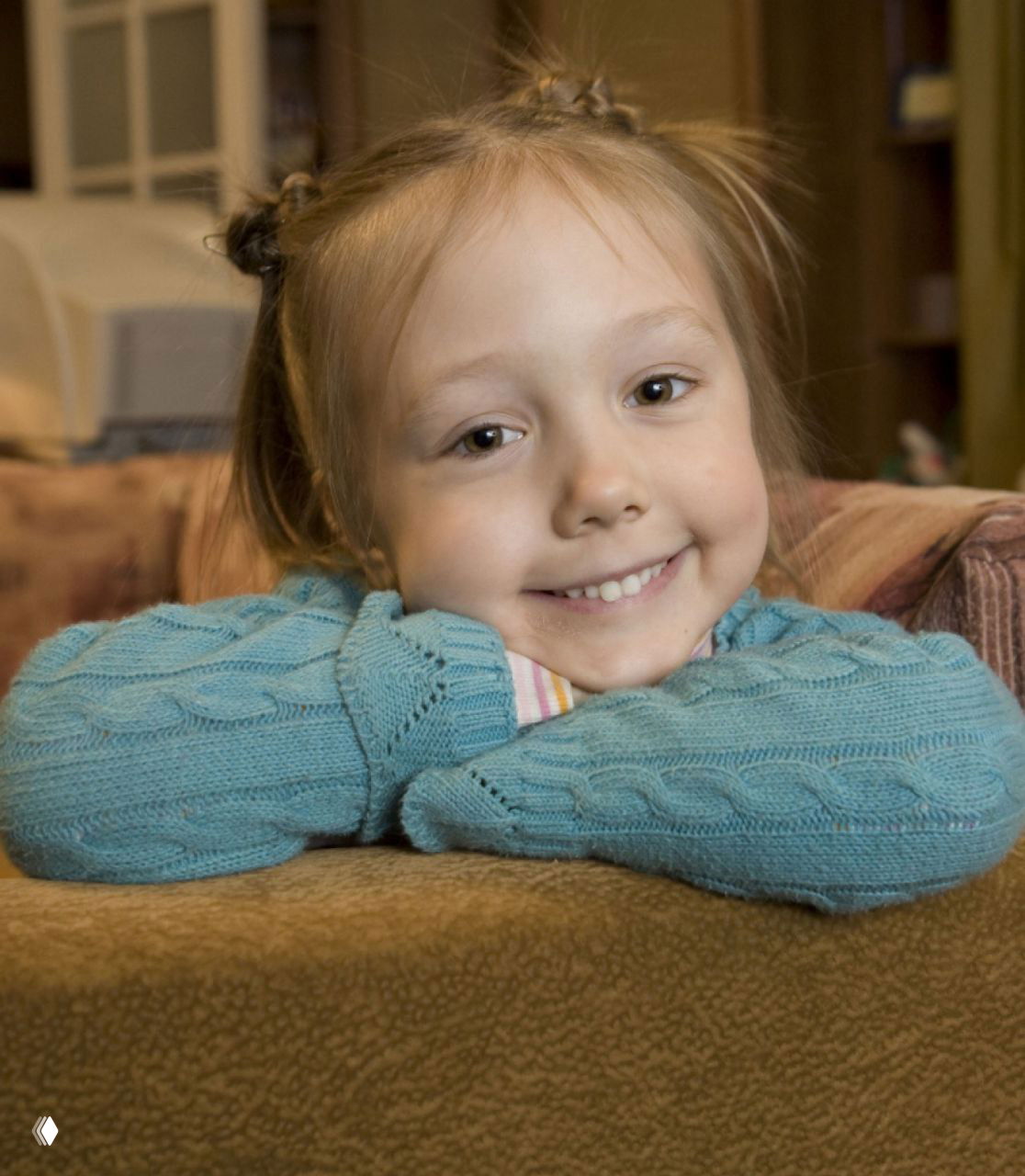 Крупный план улыбающейся маленькой актрисы в голубом свитере, она опирается на диван, тёплая домашняя обстановка и детская искренняя улыбка.