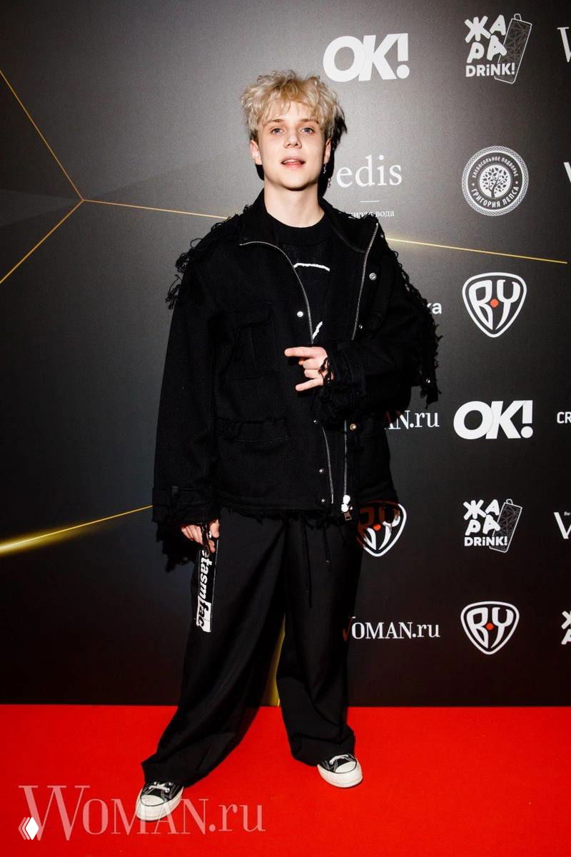 Полный рост молодого артиста в чёрной свободной одежде на красной дорожке — фото со светского мероприятия с логотипами спонсоров на фоне.