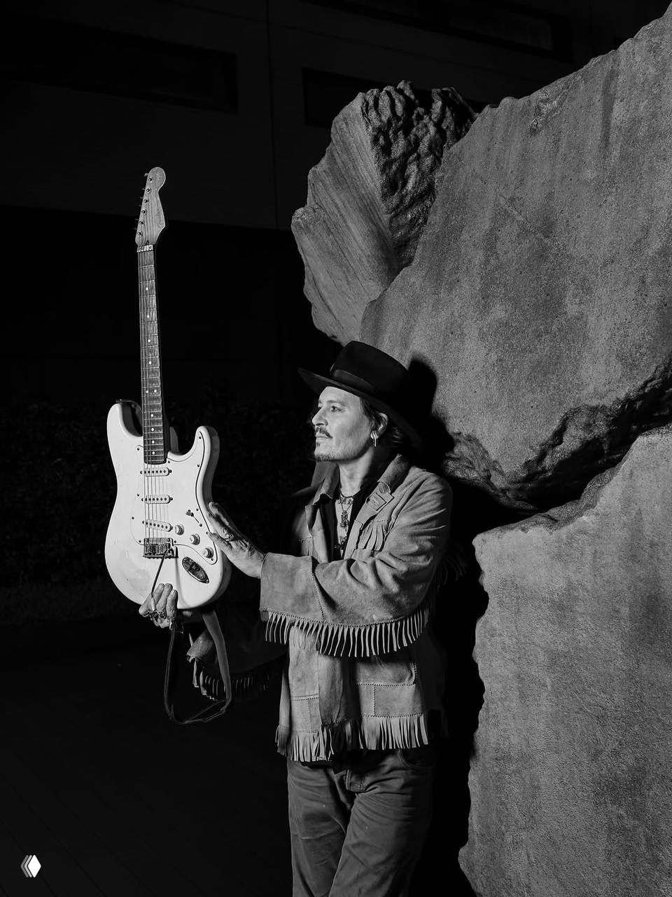 Чёрно-белый художественный кадр в Токио: мужчина в шляпе и бахромчатой куртке держит электрогитару у крупных каменных форм ночью, ретро-образ сценической съёмки