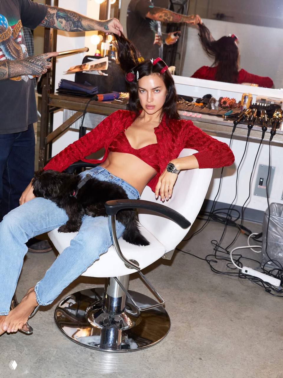 Ирина Шейк в кресле гримерки с бигуди, джинсами и красным кардиганом; кадр бэкстейджа съёмки с инструментами визажиста.