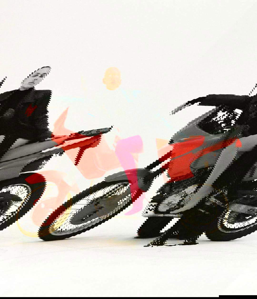 Модель позирует на красном мотоцикле на белом фоне: контраст чёрной одежды и яркой обуви, динамичный модный кадр.