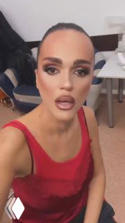 Крупный портрет женщины в красном топе с ярким макияжем, снятой в помещении в полублизком плане; видны лицо, плечи и часть интерьера с креслом и столом.