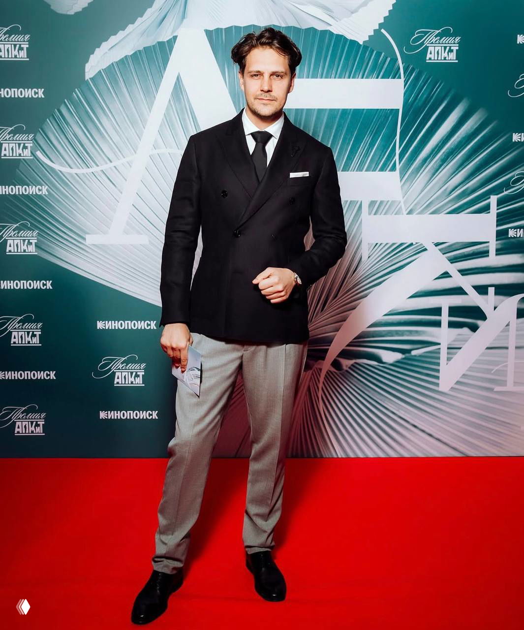 Актёр Милош Бикович на красной дорожке у прессволла, официальный наряд, поза для фотосессии и логотипы кинонаград на фоне.