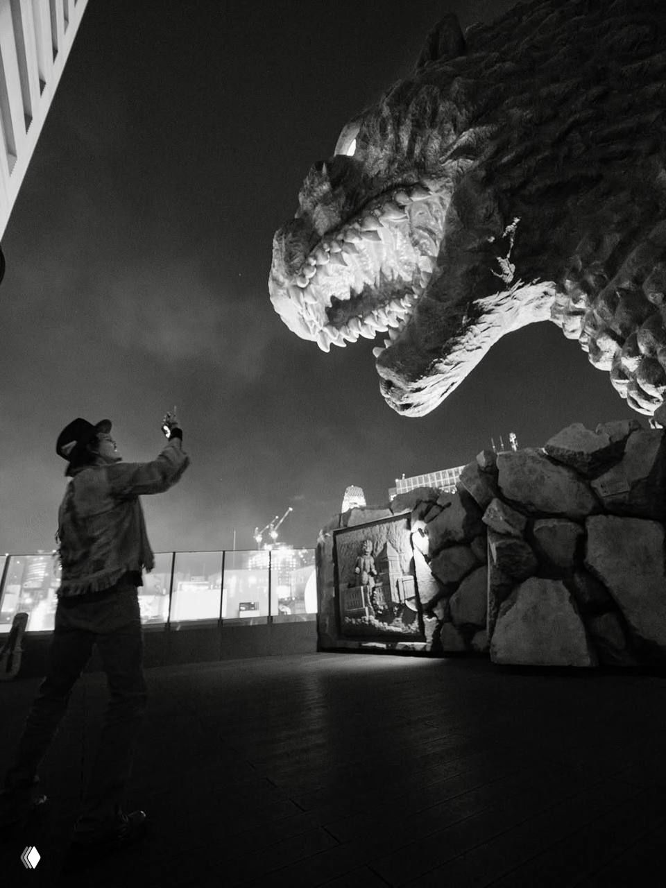 Мужчина в шляпе тянется к большой скульптуре монстра на крыше здания, ночная ч/б сцена с городскими огнями и атмосферной подсветкой, художественная постановка