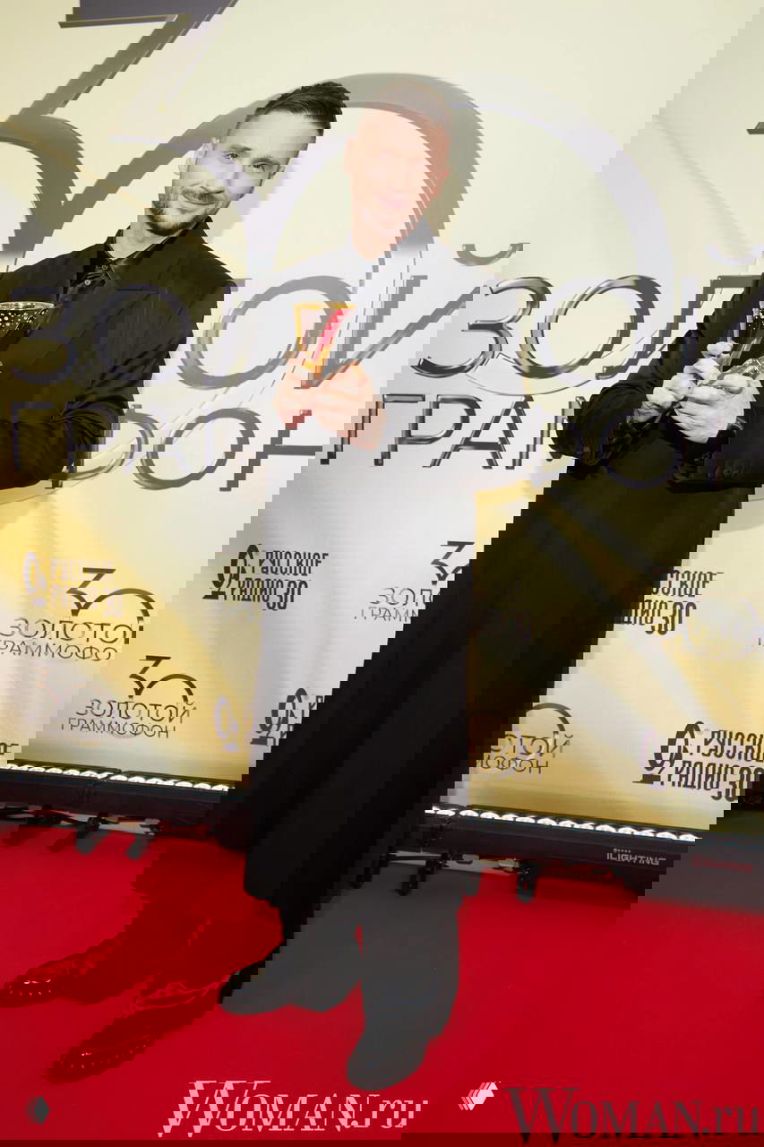 Сергей Лазарев в тёмном длинном пальто держит награду и улыбается на красной дорожке юбилейного шоу «Русское радио», видно число 30 на фоне.