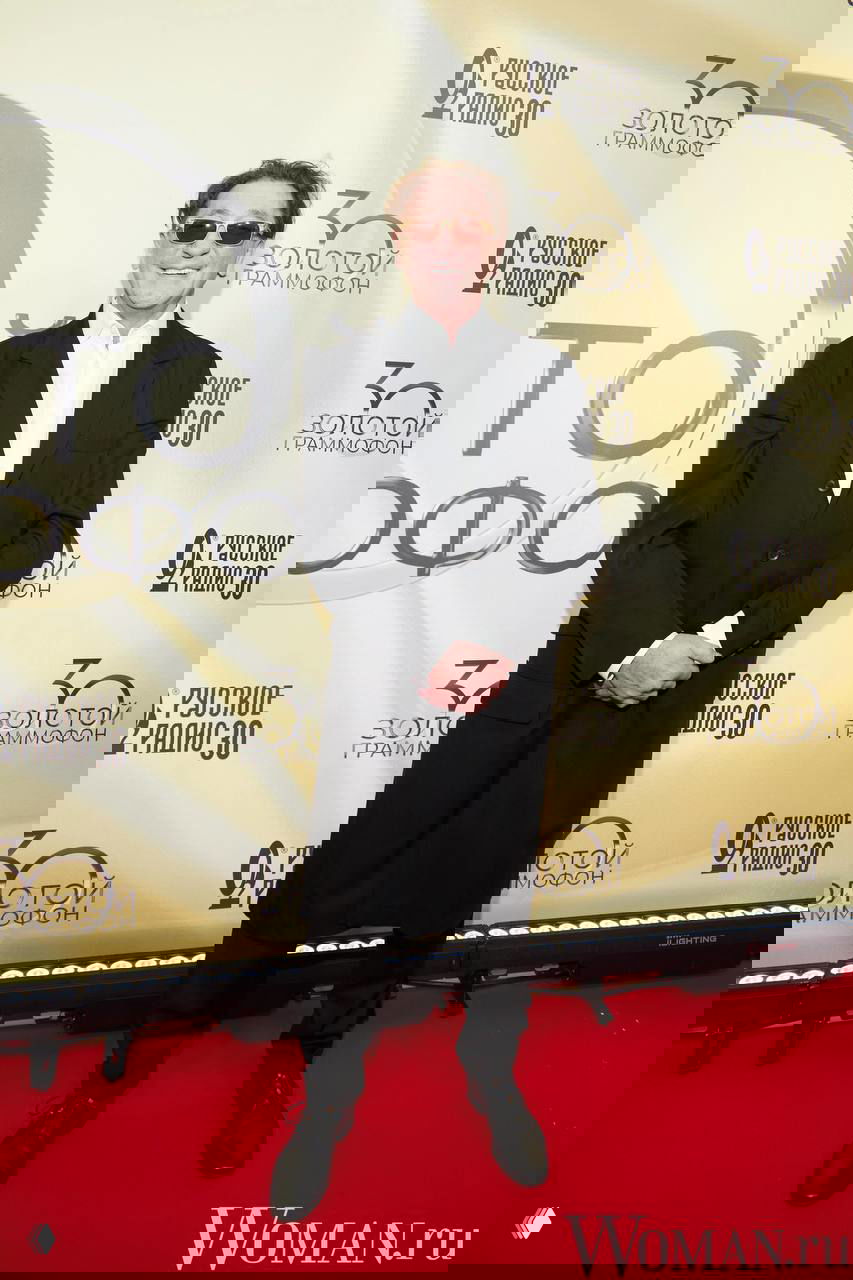 Григорий Лепс в чёрном пальто и солнцезащитных очках позирует на красной дорожке юбилейного шоу «Русское радио», логотип и декор видны на фоне.