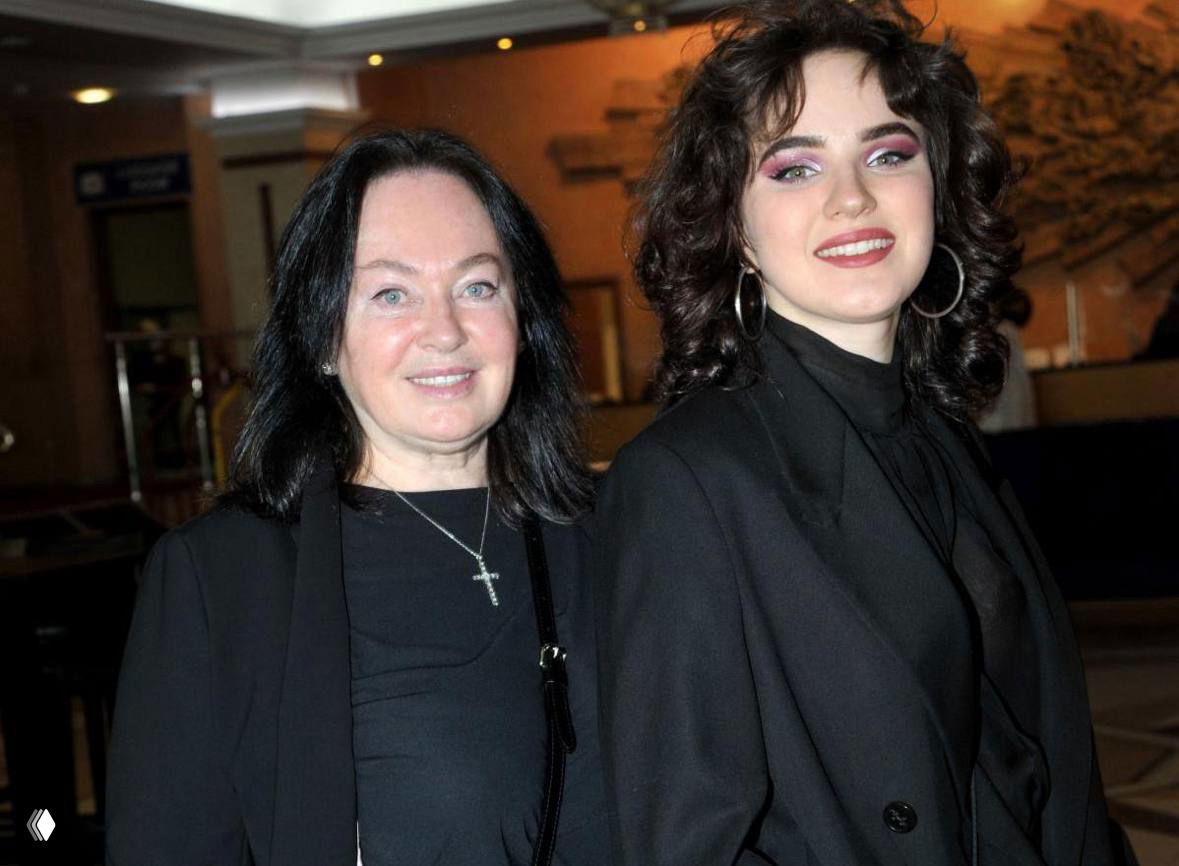 Фотография в помещении: две женщины в тёмной одежде стоят рядом; старшая улыбается, молодая — в ярком макияже и крупных серьгах.