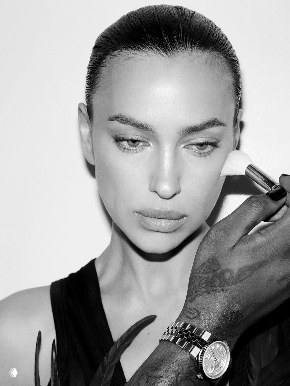 Крупный чёрно‑белый портрет Ирины Шейк при нанесении макияжа; детализированная съёмка лица, кисть визажиста рядом с щекой.