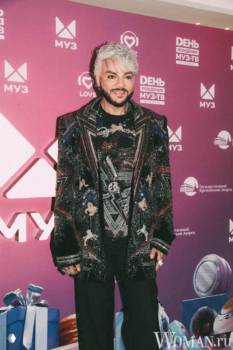 Филипп Киркоров в блестящем наряде на фоне брендированной фотозоны Кремля; портрет артиста в полный рост на мероприятии