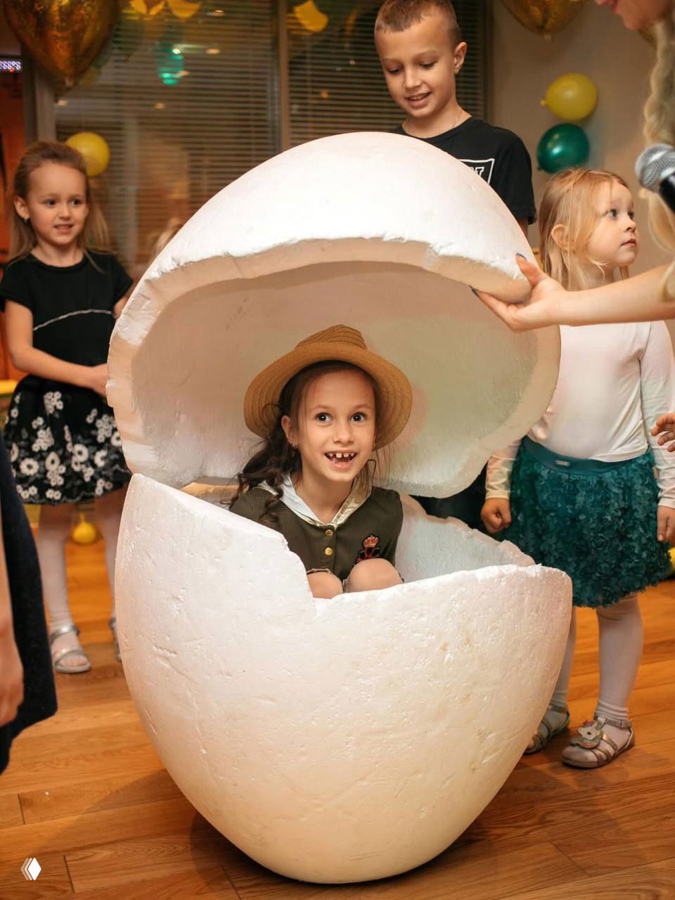 Детская вечеринка: ребенок сидит внутри большой декорации в форме разбитого яйца, вокруг другие дети и праздничный деревянный пол.