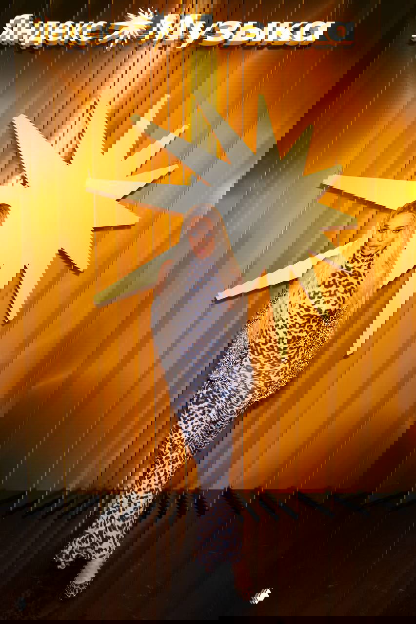 Блондинка в леопардовом платье и длинных перчатках позирует на фоне золотой инсталляции с логотипом «Яндекс Музыка» на красной дорожке.