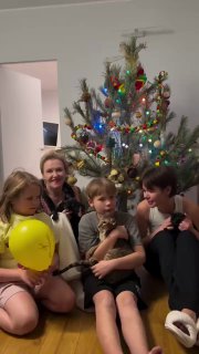 Вертикальный кадр домашнего видео: дети и взрослые сидят возле украшенной ёлки, видны игрушки и шарики, живые эмоции.