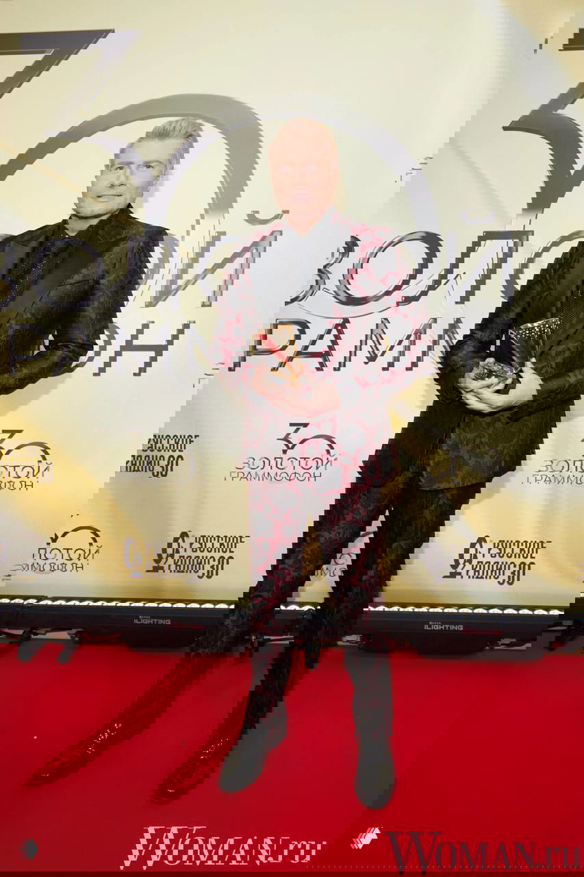 Николай Басков в тёмном костюме с узорчатой тканью и чёрной рубашке держит статуэтку на красной дорожке, фон украшен логотипами и золотым декором.