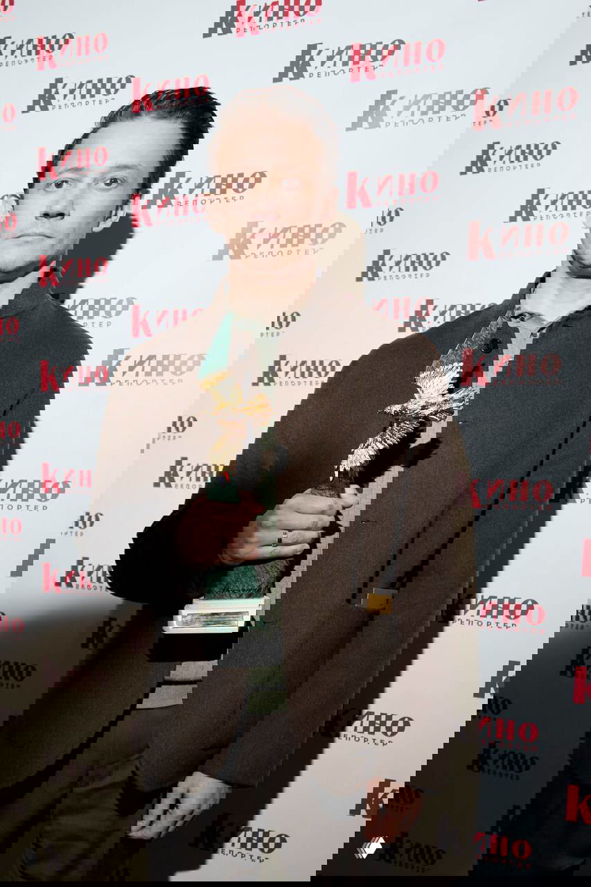 Фотография победителя с наградой, мужчина в коричневом пальто держит статуэтку, позирует на фоне пресс-волла с логотипом «Кино репортёр».