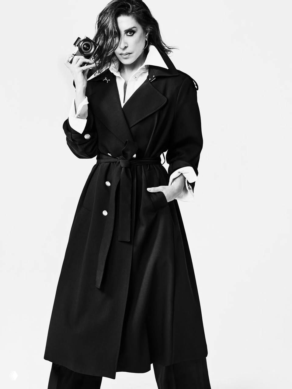 Полнофигурный чёрно‑белый кадр женщины в длинном тренче с фотоаппаратом, динамичная поза на нейтральном фоне