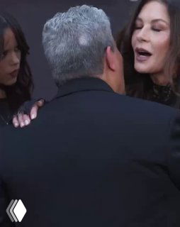 Кадр со съёмок события: Кэтрин Зета‑Джонс здоровается с Луисом Гусманом на красной дорожке, у обоих дружелюбные жесты и фон с тематикой шоу.