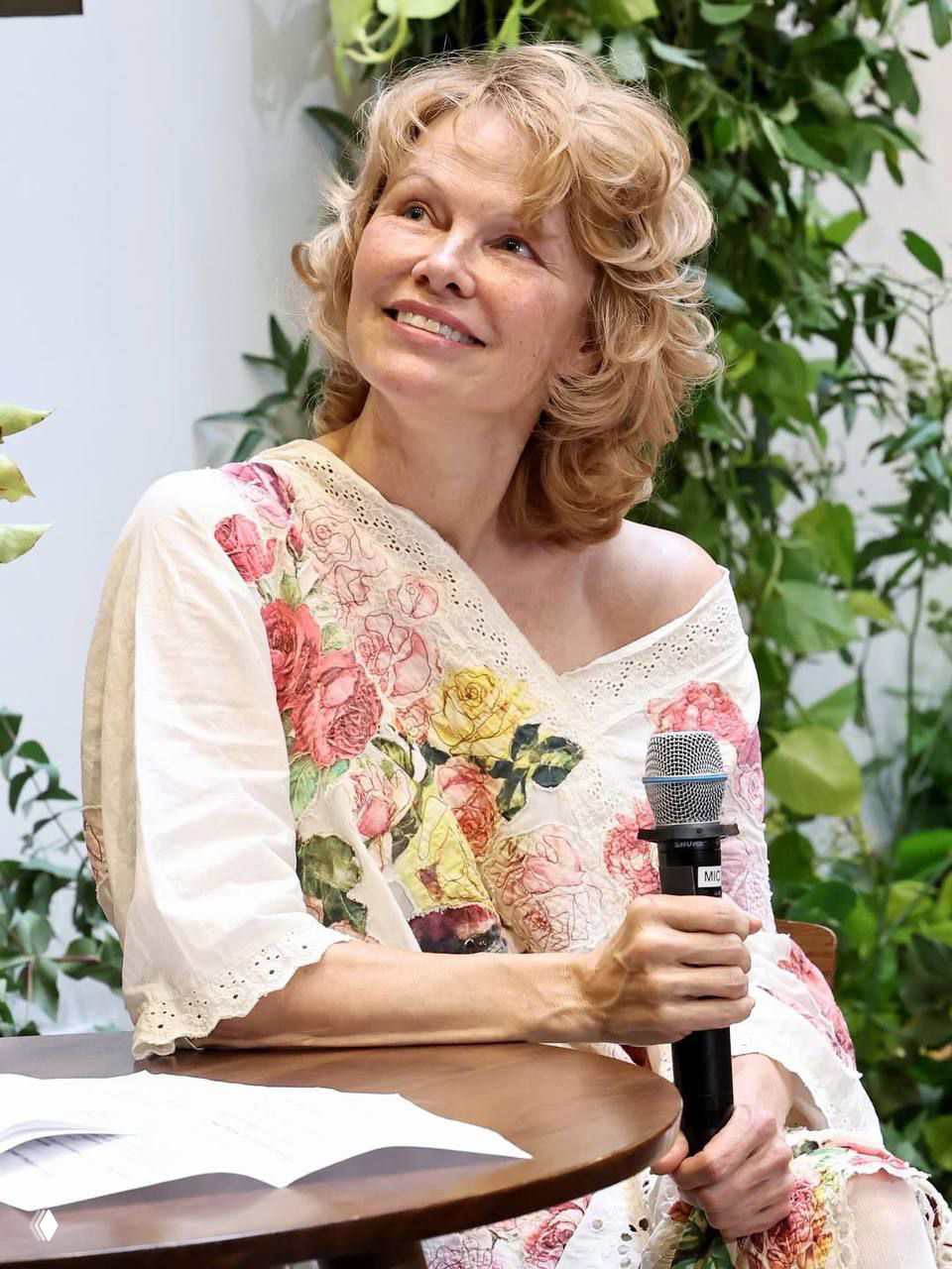 Памела Андерсон в светлом платье с розовыми и жёлтыми цветами, улыбается и держит микрофон на открытии pop-up магазина Sonsie.