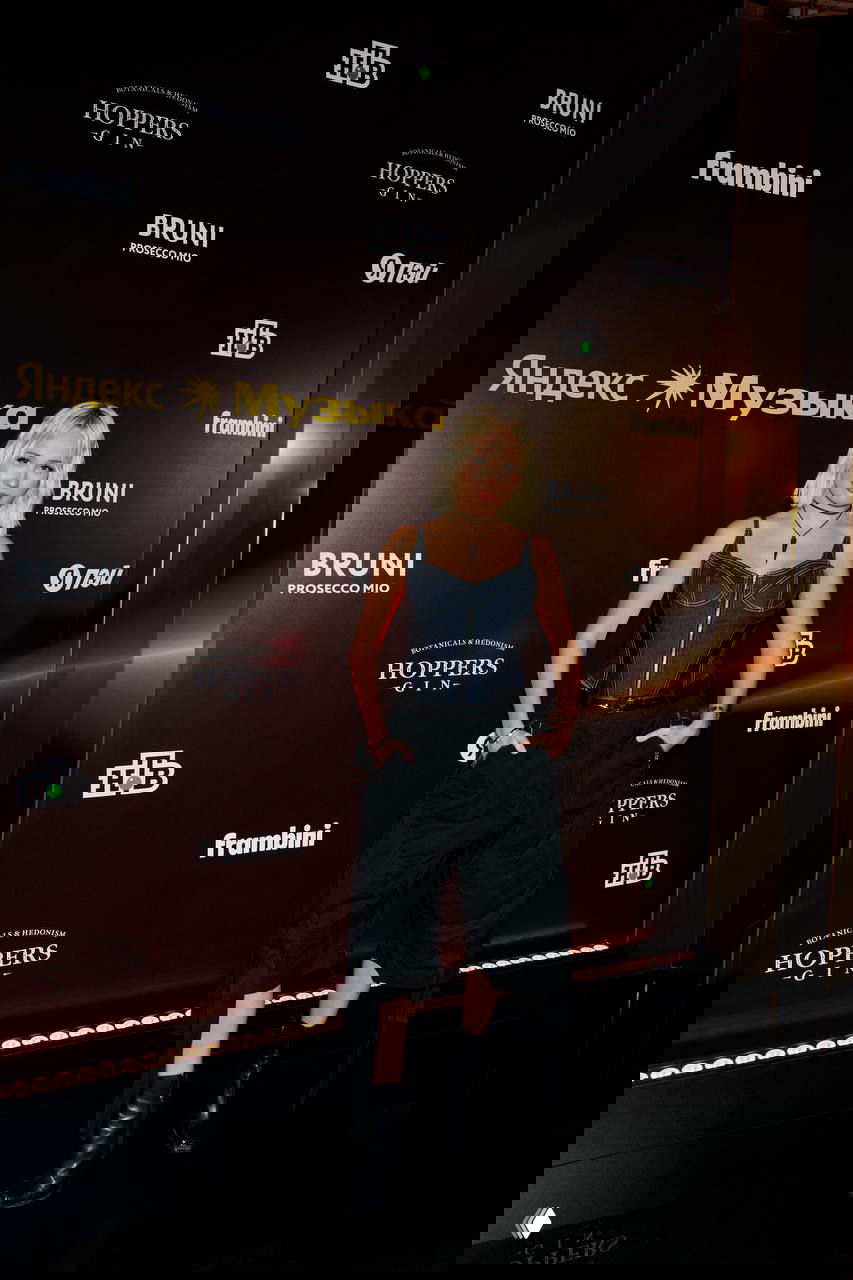 Женщина в джинсовом корсете и черных брюках позирует на пресс‑волле с логотипом «Яндекс Музыка» на премии.